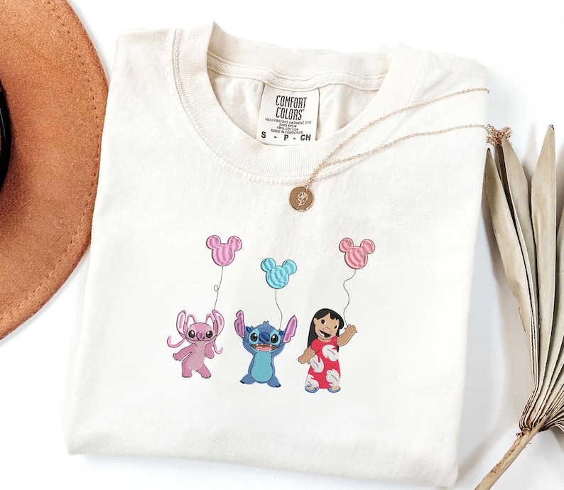 K&ouml;nnte beinhalten: Ein cremefarbenes Kurzarm-T-Shirt mit einer Stickerei von zwei blauen und rosa au&szlig;erirdischen Charakteren und einem M&auml;dchen, das drei pinke und blaue Luftballons in Mickey-Mouse-Form h&auml;lt. Das Shirt hat ein Comfort Colors Markenetikett am Hals.