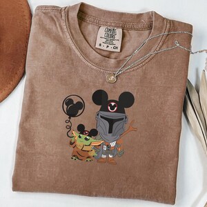 Op de afbeelding: Een bruin T-shirt met korte mouwen en een grafische print van een personage met een helm met Mickey Mouse-oren, die een ballon vasthoudt naast een klein groen wezen dat ook Mickey Mouse-oren draagt.