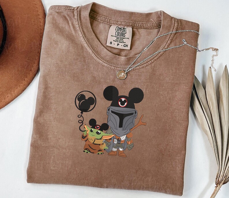 Op de afbeelding: Een bruin T-shirt met korte mouwen en een grafische print van een personage met een helm met Mickey Mouse-oren, die een ballon vasthoudt naast een klein groen wezen dat ook Mickey Mouse-oren draagt.