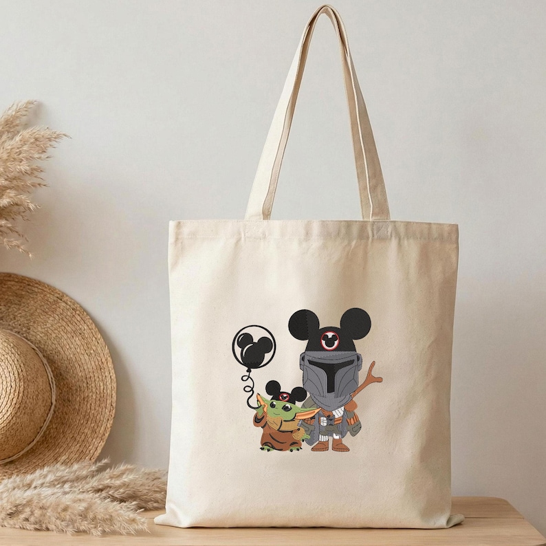 Op de afbeelding: Een natuurkleurige canvas draagtas met een grafische print van een personage met een helm en een klein groen wezen, beide met Mickey Mouse-oren. Het personage houdt een ballon vast in de vorm van Mickey Mouse-oren aan een touwtje.