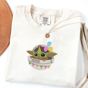 Puede incluir: Una camiseta de color crema con un gr&aacute;fico de una criatura verde de ojos grandes que lleva un gorro de fiesta amarillo con dibujos. La criatura est&aacute; dentro de una c&aacute;psula circular gris decorada con globos de colores y una guirnalda de banderines.