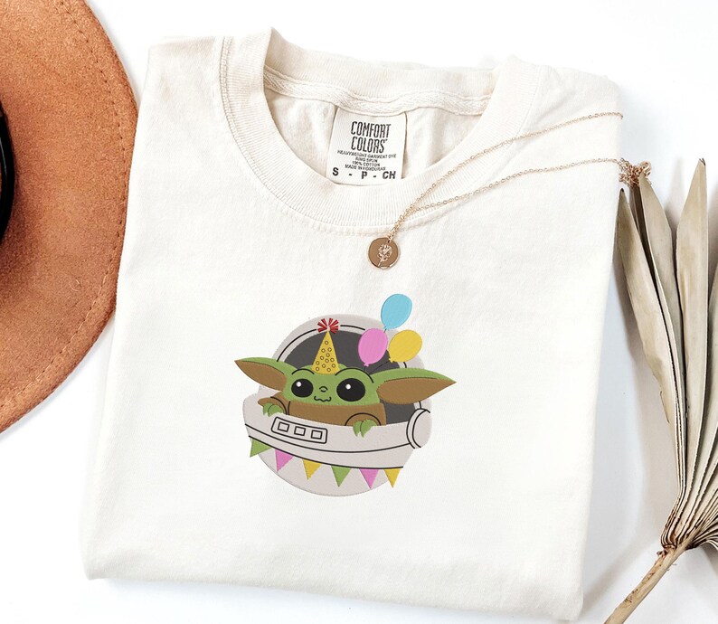 Puede incluir: Una camiseta de color crema con un gr&aacute;fico de una criatura verde de ojos grandes que lleva un gorro de fiesta amarillo con dibujos. La criatura est&aacute; dentro de una c&aacute;psula circular gris decorada con globos de colores y una guirnalda de banderines.