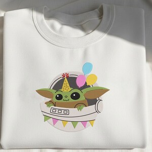 Puede incluir: Una sudadera blanca de cuello redondo con un bordado de una criatura verde de orejas grandes que lleva un gorro de fiesta amarillo con puntos rojos. La criatura est&aacute; dentro de un casco espacial blanco con globos rosas, azules y amarillos.