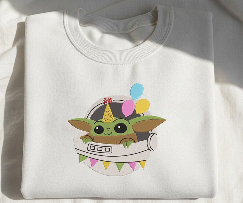 Puede incluir: Una sudadera blanca de cuello redondo con un bordado de una criatura verde de orejas grandes que lleva un gorro de fiesta amarillo con puntos rojos. La criatura est&aacute; dentro de un casco espacial blanco con globos rosas, azules y amarillos.