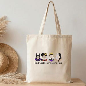 May include: Beige canvas tote bag featuring a graphic of four stylized female figures with dark hair and purple accents. The text "Bad Girls Have More Fun" is printed below the figures. The bag has long handles.