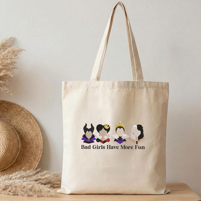 May include: Beige canvas tote bag featuring a graphic of four stylized female figures with dark hair and purple accents. The text "Bad Girls Have More Fun" is printed below the figures. The bag has long handles.