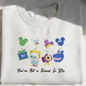 May include: A white crewneck sweatshirt featuring an embroidered design of four colorful characters with spring-like attachments and the text "You've Got a Friend In Me" printed in black script below the graphic elements on the front of the garment.