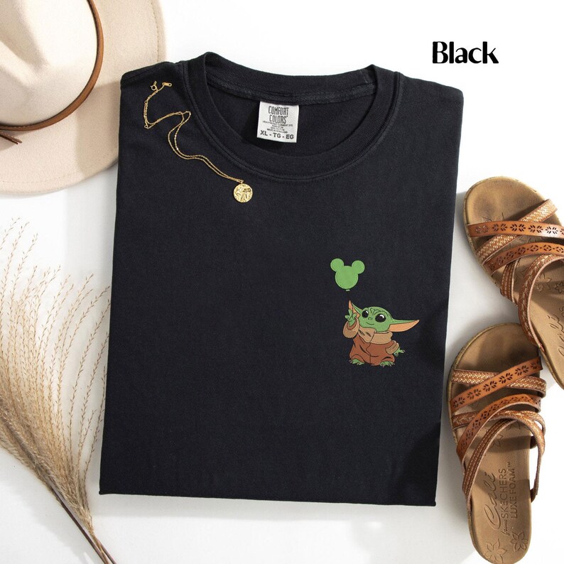 May include: A black crew neck t-shirt featuring a small embroidered graphic of a green creature in a brown robe holding a green Mickey Mouse-shaped balloon. The shirt has a Comfort Colors brand label at the inner neckline and is labeled as size XL.