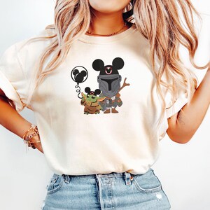 Op de afbeelding: Een cr&egrave;mekleurig T-shirt met korte mouwen en een grafische print van een ruimte-premiejager en een klein groen wezen, beide met Mickey Mouse-oren op. Het kleine wezen houdt een zwarte ballon in de vorm van Mickey Mouse vast.
