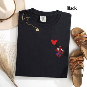May include: Black crew neck t-shirt with an embroidered Spider-Man character holding a Mickey Mouse balloon. The shirt also features a gold necklace and is paired with brown sandals and a beige hat.