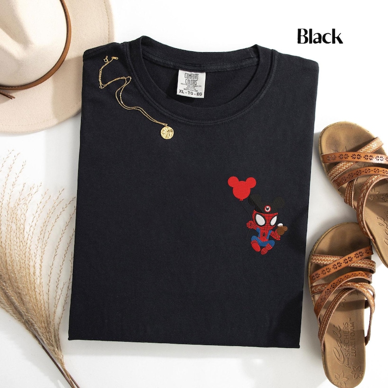 May include: Black crew neck t-shirt with an embroidered Spider-Man character holding a Mickey Mouse balloon. The shirt also features a gold necklace and is paired with brown sandals and a beige hat.