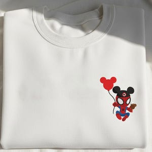 May include: White sweatshirt featuring a cartoon character mashup of Spider-Man and Mickey Mouse. The character is holding a red balloon and a small brown object. The design is in red, blue, black, and brown.