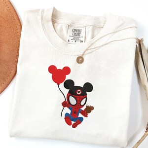 May include: Cream-colored t-shirt featuring an embroidered cartoon character combining Mickey Mouse and Spider-Man. The character holds a red balloon and a small treat. A gold necklace with a pendant is draped across the shirt.