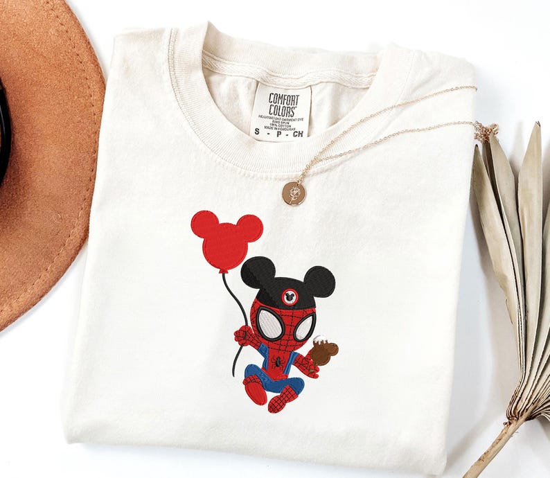 May include: Cream-colored t-shirt featuring an embroidered cartoon character combining Mickey Mouse and Spider-Man. The character holds a red balloon and a small treat. A gold necklace with a pendant is draped across the shirt.