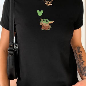 May include: A black short-sleeved t-shirt featuring a graphic of a small green creature with large ears wearing a brown robe. The creature is holding a light green balloon shaped like a mouse head. The shirt has a crew neckline and a simple fit.