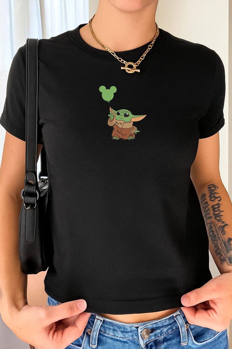 May include: A black short-sleeved t-shirt featuring a graphic of a small green creature with large ears wearing a brown robe. The creature is holding a light green balloon shaped like a mouse head. The shirt has a crew neckline and a simple fit.
