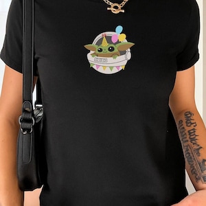 Puede incluir: Una camiseta negra de manga corta con el gr&aacute;fico de una peque&ntilde;a criatura verde de ojos grandes que lleva un gorro de fiesta colorido dentro de una c&aacute;psula espacial blanca. La c&aacute;psula est&aacute; decorada con banderines y tres globos flotantes.