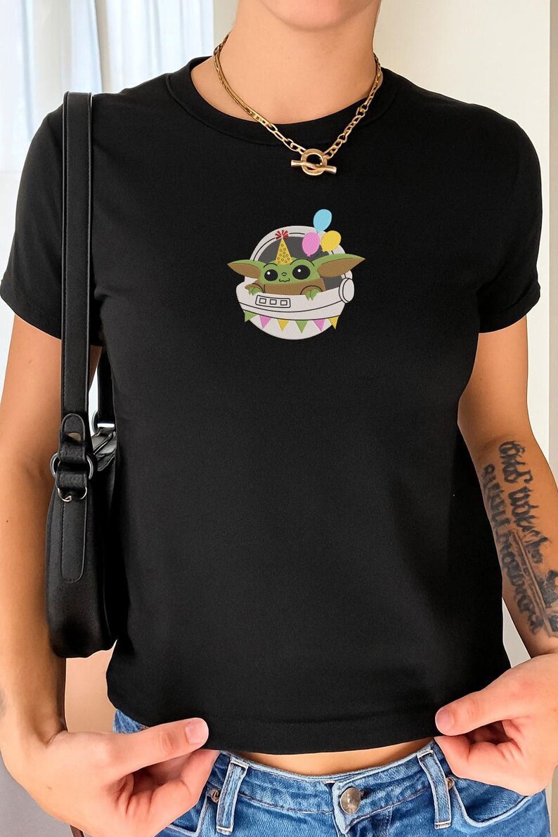 Puede incluir: Una camiseta negra de manga corta con el gr&aacute;fico de una peque&ntilde;a criatura verde de ojos grandes que lleva un gorro de fiesta colorido dentro de una c&aacute;psula espacial blanca. La c&aacute;psula est&aacute; decorada con banderines y tres globos flotantes.
