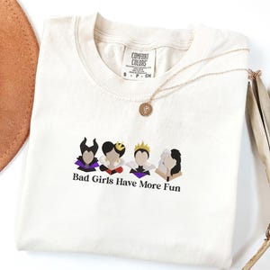 May include: Cream-colored t-shirt with the text "Bad Girls Have More Fun" and illustrations of four cartoon characters. A gold necklace with a pendant is draped across the shirt. A brown hat and dried plant are in the background.