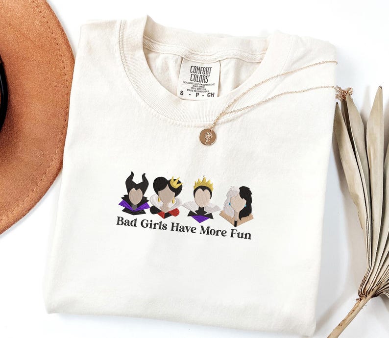 May include: Cream-colored t-shirt with the text "Bad Girls Have More Fun" and illustrations of four cartoon characters. A gold necklace with a pendant is draped across the shirt. A brown hat and dried plant are in the background.