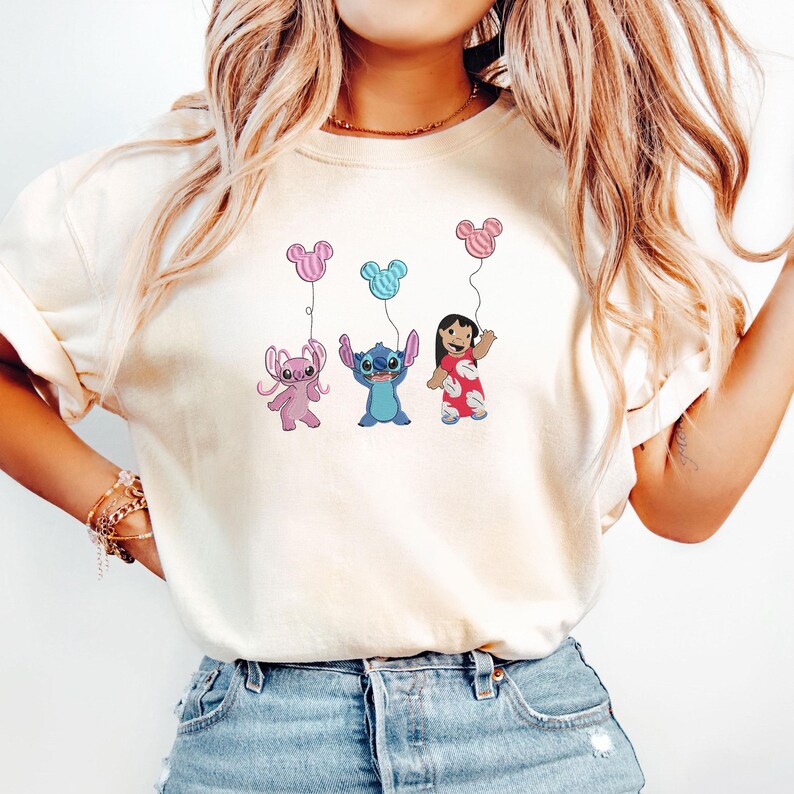 K&ouml;nnte beinhalten: Ein cremefarbenes T-Shirt mit einem Grafikdruck von Angel, Stitch und Lilo aus Lilo und Stitch. Jede Figur h&auml;lt einen Luftballon in Form eines Mickey-Maus-Kopfes in Rosa, Blau oder Rot, passend zu den jeweiligen Farben und Outfits.