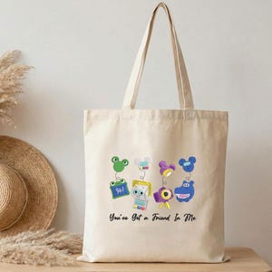 May include: A beige canvas tote bag featuring a graphic print of four colorful cartoon characters connected by springs to floating heads. Below the characters, the black text reads, You've Got a Friend In Me. The bag has two long shoulder straps.