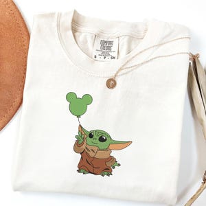 May include: An off-white short-sleeve t-shirt featuring an embroidered graphic of a small green creature with large ears wearing a brown robe and holding a green balloon shaped like a mouse head. The shirt has a Comfort Colors brand neck label.