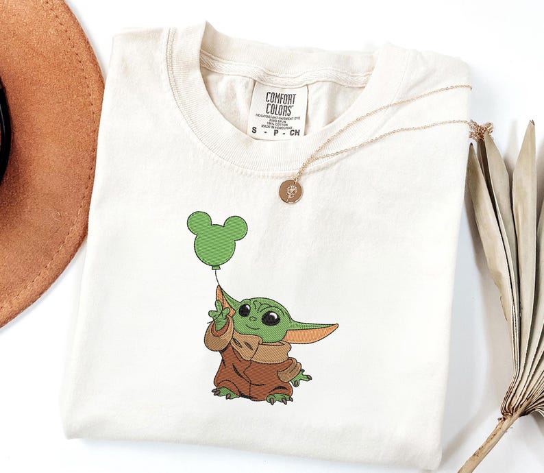 May include: An off-white short-sleeve t-shirt featuring an embroidered graphic of a small green creature with large ears wearing a brown robe and holding a green balloon shaped like a mouse head. The shirt has a Comfort Colors brand neck label.