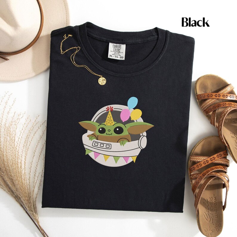 Puede incluir: Una camiseta negra con el gr&aacute;fico de una criatura verde de ojos grandes que lleva un gorro de fiesta amarillo con puntos rojos. La criatura est&aacute; dentro de una c&aacute;psula espacial blanca decorada con globos de colores y una guirnalda triangular.