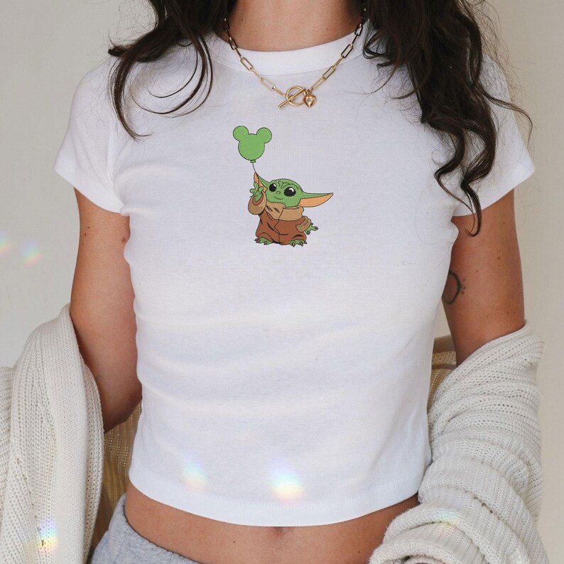 May include: A white short-sleeved t-shirt featuring a graphic of a small green creature with large ears wearing a brown robe and holding a green balloon shaped like three circles. The shirt is paired with a gold-toned chain link necklace.