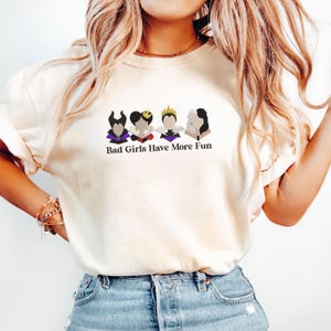 May include: Cream-colored t-shirt featuring a graphic of four cartoon villain characters with the text "Bad Girls Have More Fun". The graphic includes characters with horns, crowns, and stylized hair. The shirt is paired with blue denim jeans.