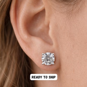 May include: A close-up of a sparkling, round-cut diamond stud earring set in a silver or white gold setting. The earring is displayed on an ear, with the text "READY TO SHIP" in a white rectangle.