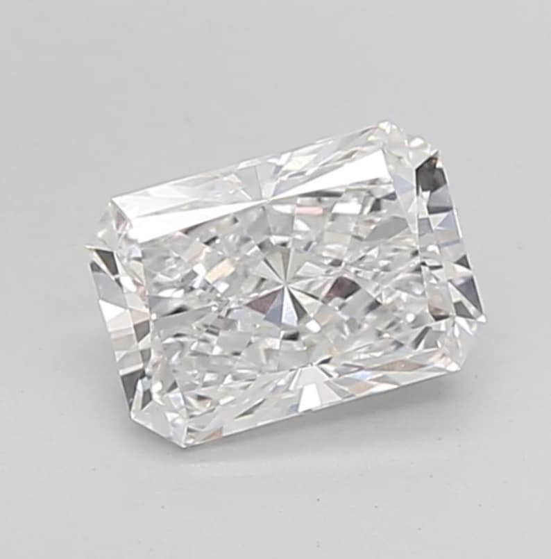 May include: A clear, radiant-cut diamond with a rectangular shape. The gemstone has many facets that reflect light. The diamond is set against a plain, light background.