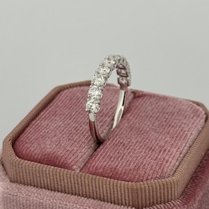 Puede incluir: Un anillo de plata con una fila de piedras preciosas redondas y transparentes. El anillo est&aacute; colocado sobre un joyero de terciopelo rosa. Las piedras preciosas est&aacute;n espaciadas uniformemente y brillan. La banda del anillo es delgada y pulida.