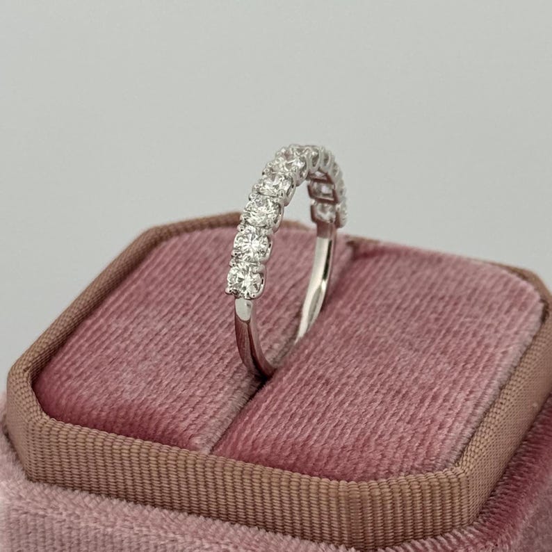 Puede incluir: Un anillo de plata con una fila de piedras preciosas redondas y transparentes. El anillo est&aacute; colocado sobre un joyero de terciopelo rosa. Las piedras preciosas est&aacute;n espaciadas uniformemente y brillan. La banda del anillo es delgada y pulida.