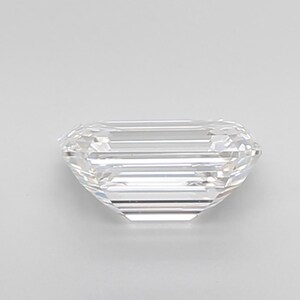 May include: A clear, emerald-cut gemstone with a rectangular shape and faceted surfaces. The gemstone is colorless and appears to be highly reflective, with visible internal structures. The facets create a play of light and shadow, enhancing its brilliance.