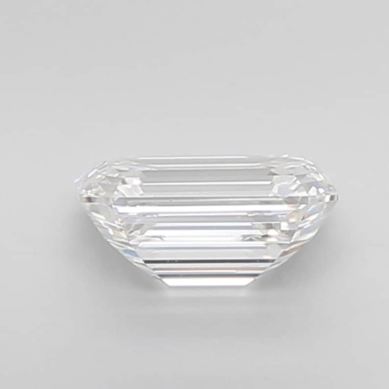 May include: A clear, emerald-cut gemstone with a rectangular shape and faceted surfaces. The gemstone is colorless and appears to be highly reflective, with visible internal structures. The facets create a play of light and shadow, enhancing its brilliance.