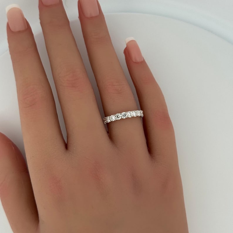 Puede incluir: Primer plano de una mano con un anillo de diamantes de la eternidad. El anillo presenta una fila continua de diamantes de talla redonda engastados en una banda de metal blanco. Los diamantes brillan, reflejando la luz. Es una joya cl&aacute;sica.