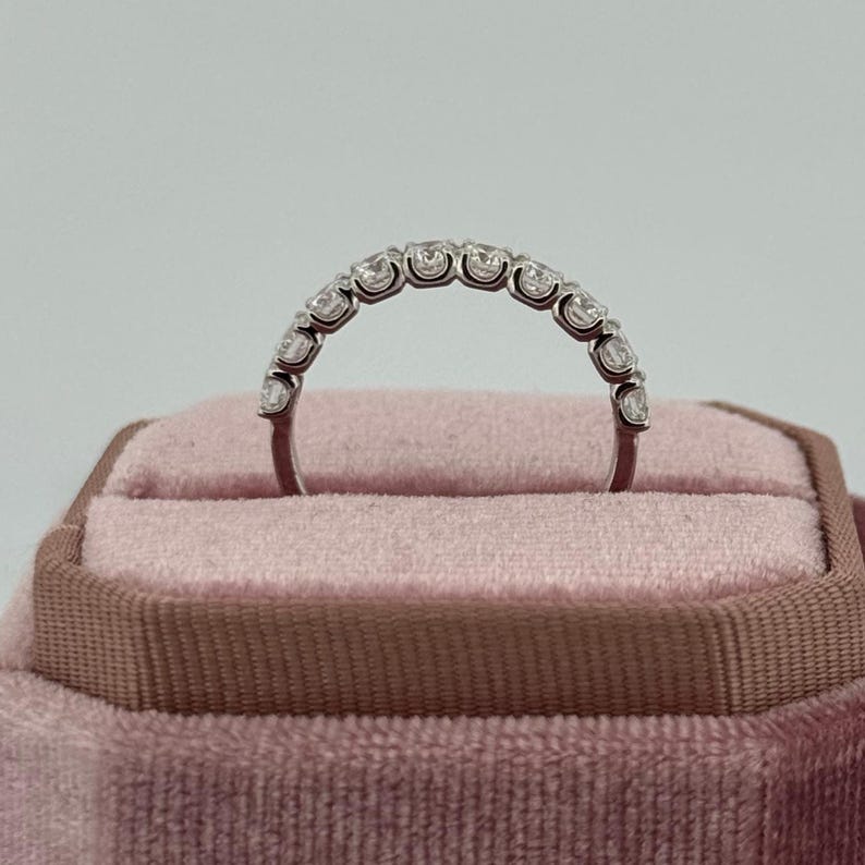 Puede incluir: Un anillo de plata con una fila de piedras preciosas redondas y transparentes. El anillo se muestra sobre un fondo de terciopelo rosa. Las piedras est&aacute;n espaciadas uniformemente, formando un arco.