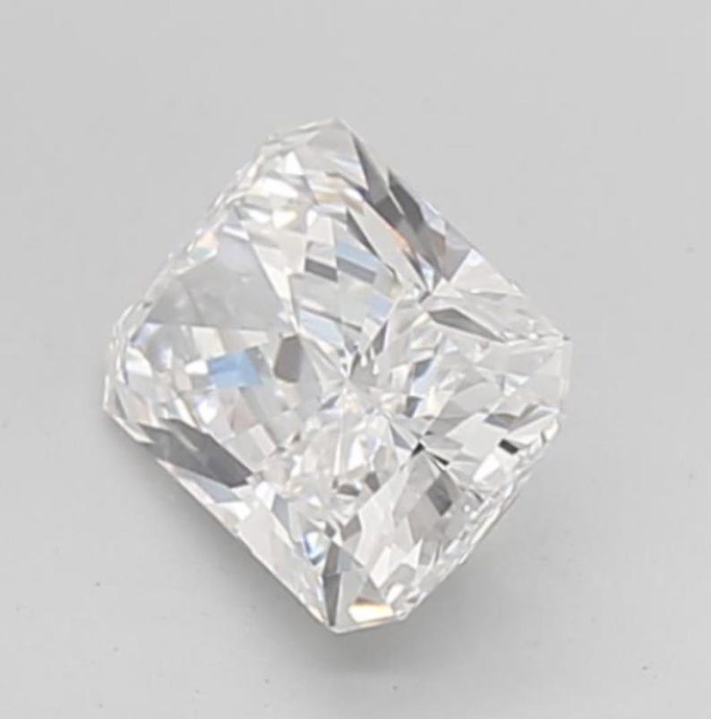 May include: A square-shaped, radiant-cut diamond with multiple facets, reflecting light. The colorless gemstone sparkles brightly. The diamond is set against a plain, light background.