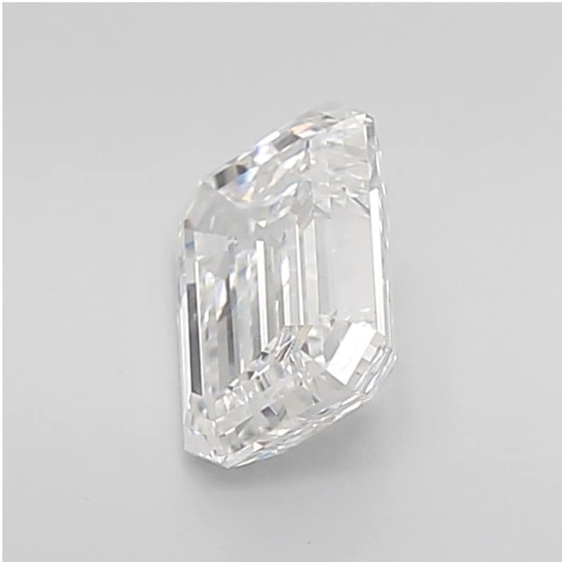 May include: A clear, emerald-cut diamond with a rectangular shape and beveled corners. The facets reflect light, creating a brilliant sparkle. The diamond is set against a plain white background, highlighting its clarity and brilliance.