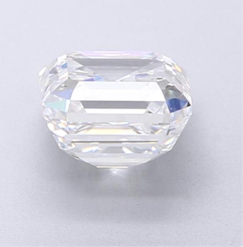 May include: A clear, faceted gemstone with an emerald cut. The gem has a rectangular shape with cut corners and a brilliant, reflective surface. The facets create a play of light and shadow, giving the stone a sparkling appearance.