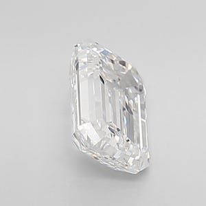 May include: A clear, emerald-cut diamond with multiple facets. The diamond is positioned at an angle, showcasing its rectangular shape and brilliant sparkle. The gemstone is set against a plain white background.