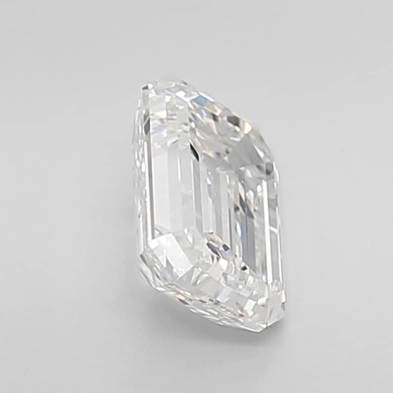 May include: A clear, emerald-cut diamond with multiple facets. The diamond is positioned at an angle, showcasing its rectangular shape and brilliant sparkle. The gemstone is set against a plain white background.