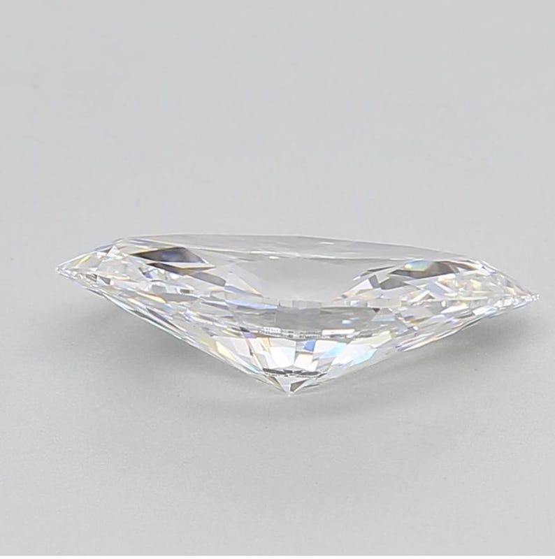 May include: A clear, faceted gemstone with a marquise cut. The diamond is highly reflective, with multiple facets catching the light. The gemstone is set against a plain, light background.