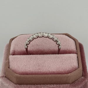 Puede incluir: Un anillo plateado con una fila de peque&ntilde;as piedras preciosas transparentes engastadas en una banda curva. El anillo se muestra en un joyero de terciopelo rosa con ribete marr&oacute;n. El fondo es de color claro y neutro.