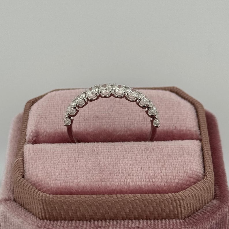 Puede incluir: Un anillo plateado con una fila de peque&ntilde;as piedras preciosas transparentes engastadas en una banda curva. El anillo se muestra en un joyero de terciopelo rosa con ribete marr&oacute;n. El fondo es de color claro y neutro.