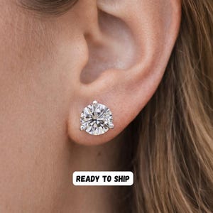 May include: A close-up of a sparkling, round-cut diamond stud earring. The earring is set in a silver or white gold setting and is worn on an ear. The image includes the text "READY TO SHIP".