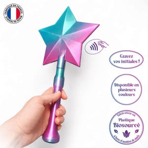 May include: A star-shaped wand in shades of blue, teal, and pink. The wand is held by a person. The image includes French text: "Made in France", "Engrave your initials!", "Available in several colours", and "Made from bio-sourced plastic".