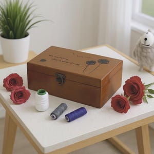 May include: A brown wooden box with a metal clasp, decorated with dandelion illustrations and text. Red roses and spools of white, grey, and blue thread are arranged around the box. A small plant and a sheep ornament are in the background.